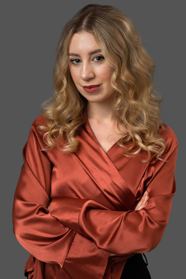 Portré egy vállig érő szőke fürtökkel rendelkező nőről, rozsdabarna szatén blúzban, keresztbe tett karral, a kamerába nézve.