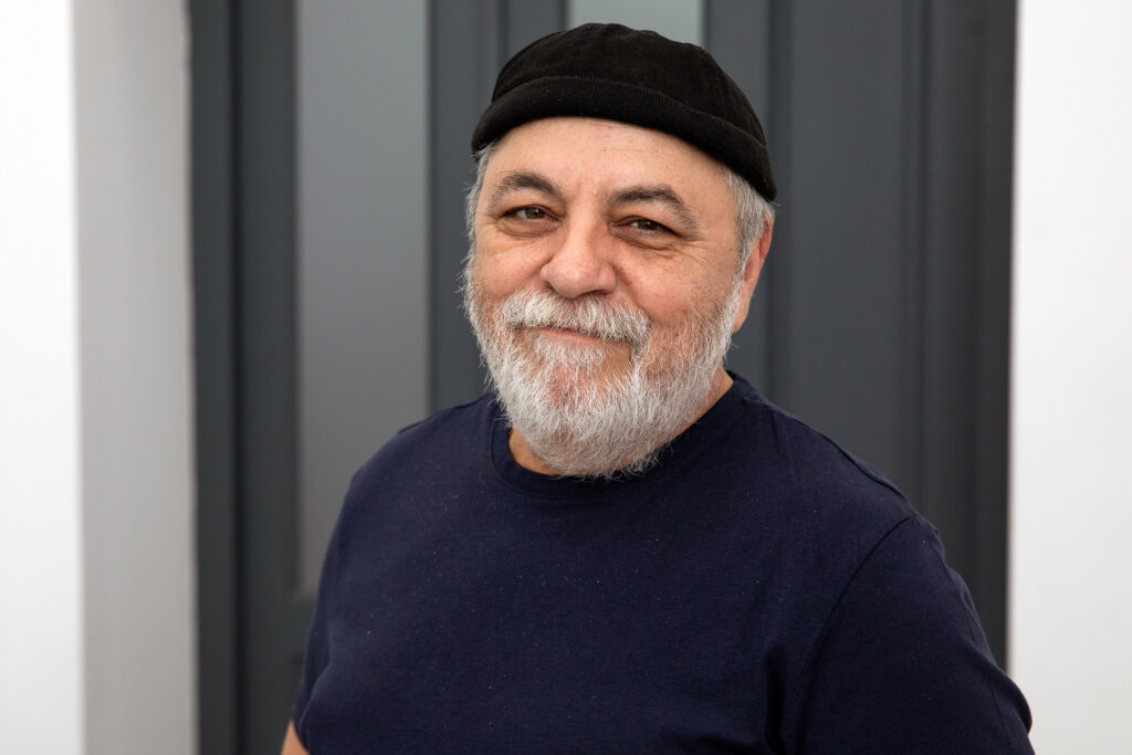 Petitoarele - repetitie 157 - Teatrul Regina Maria Oradea Smiling man with gray beard wearing a black beanie and navy shirt, standing outdoors against a grey panel background.