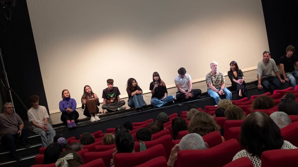 WhatsApp Image 2026-06-22 at 121115 - Teatrul Regina Maria Oradea Panel of diverse speakers seated cross-legged along a stage, one person speaking into a microphone, audience visible in red seats below.