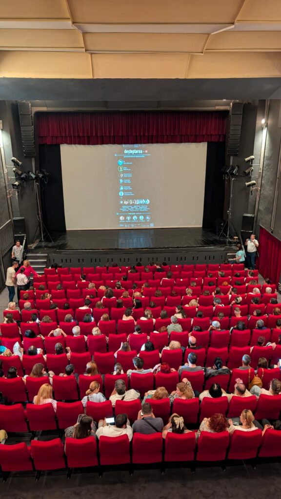 WhatsApp Image 2026-06-22 at 121124 - Teatrul Regina Maria Oradea Theater auditorium filled with rows of red seats facing a stage with a large projection screen showing a blue poster.