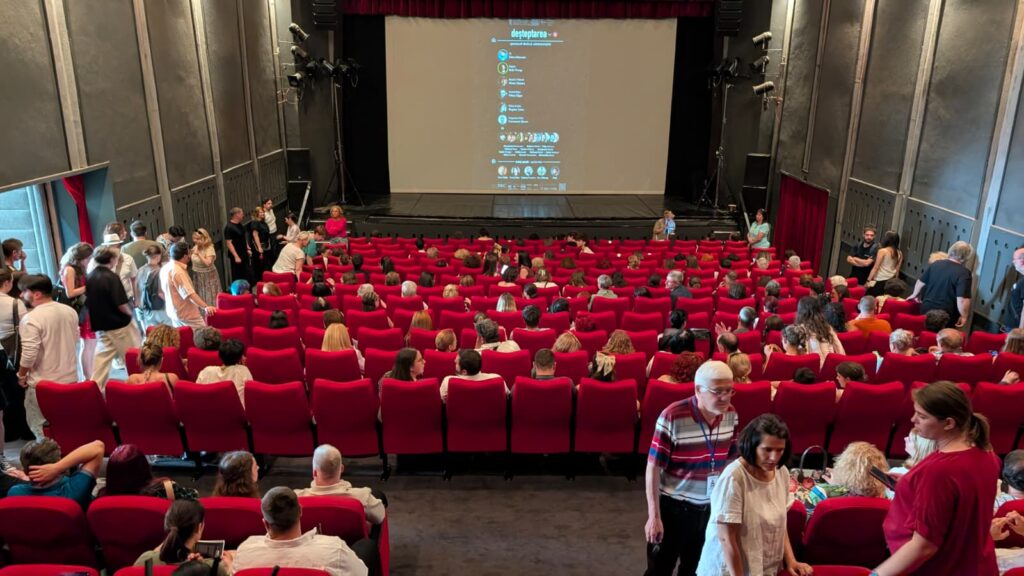 WhatsApp Image 2026-06-22 at 121313 - Teatrul Regina Maria Oradea Theater auditorium filled with people in red seats facing a blank projection screen on stage