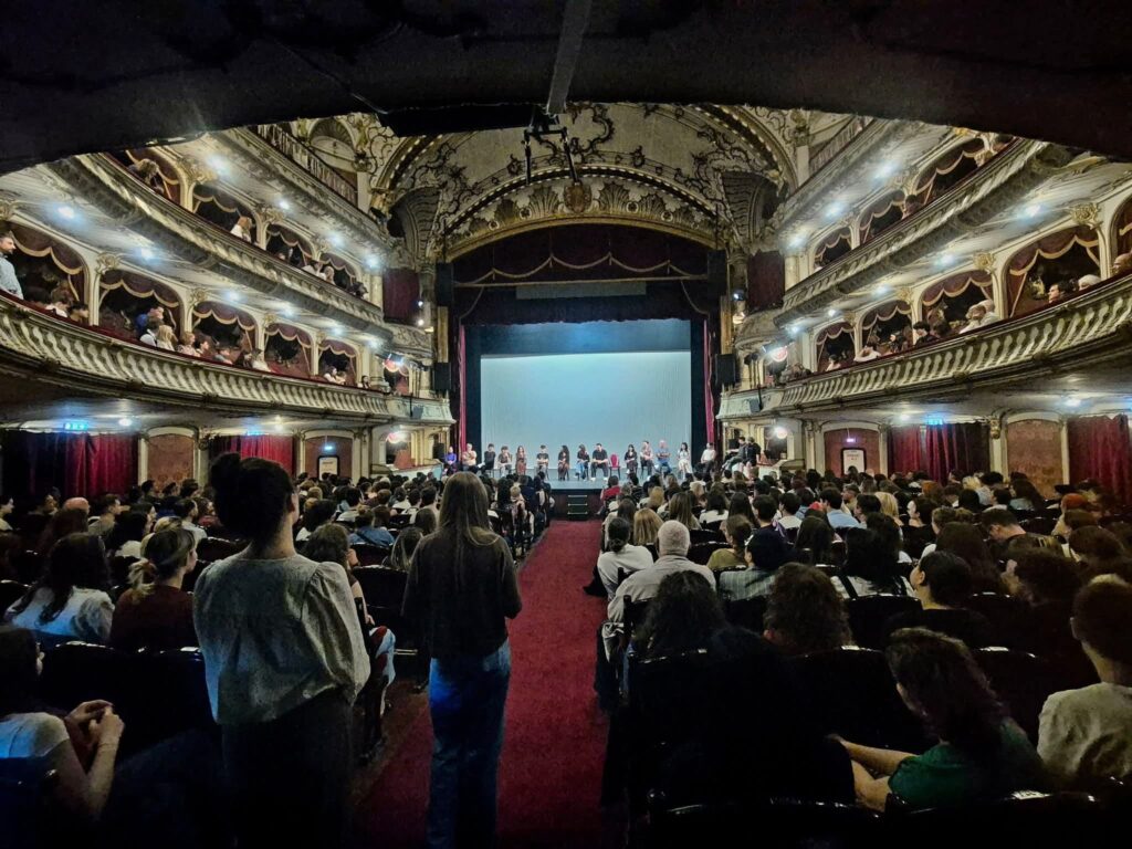 WhatsApp Image 2026-06-22 at 121332 - Teatrul Regina Maria Oradea Interior of a grand ornate theater with multiple balcony tiers filled with audience, stage at center with blue backdrop and performers lined up on stage.