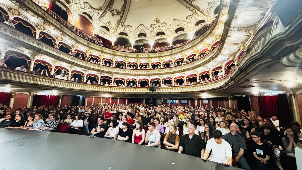 WhatsApp Image 2026-06-22 at 121403 - Teatrul Regina Maria Oradea Ornate multi-tier theater filled with seated audience facing a dark stage, gold decorations and red velvet curtains abound.