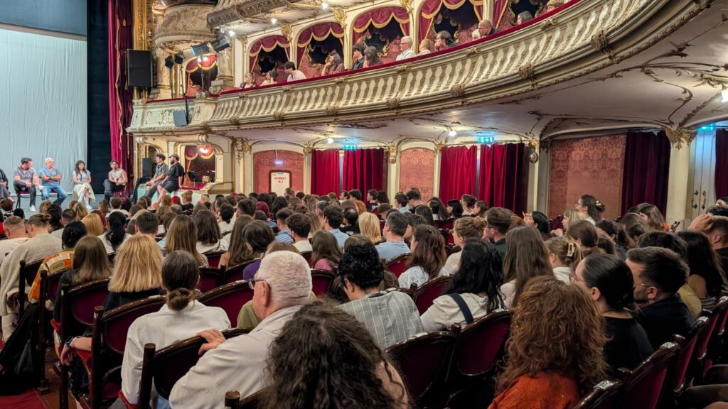 WhatsApp Image 2026-06-22 at 121529 - Teatrul Regina Maria Oradea Crowded theater with ornate gold balconies and a panel of speakers on a light stage to the left, audience watching intently.