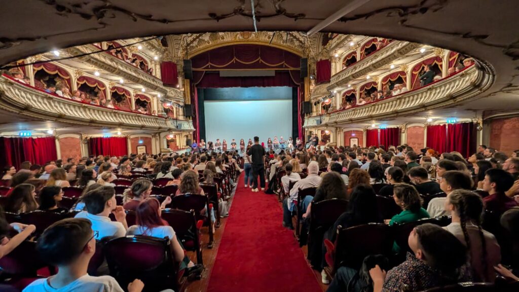 WhatsApp Image 2026-06-22 at 121555 - Teatrul Regina Maria Oradea Audience fills a ornate theater, facing a stage where a line of performers stands beneath a blue backdrop on a red-carpet aisle. Balcony boxes with gold detailing surround the scene.