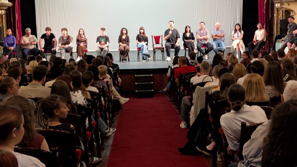 WhatsApp Image 2026-06-22 at 121610 2 - Teatrul Regina Maria Oradea Panel of speakers seated in a row on stage addressing a large audience in a theater theater.