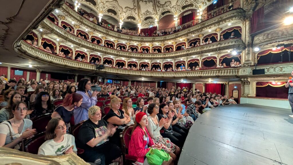 WhatsApp Image 2026-06-22 at 121611 1 - Teatrul Regina Maria Oradea Full view of a packed, ornate theater with multiple gilded balconies and red velvet seats facing the stage.