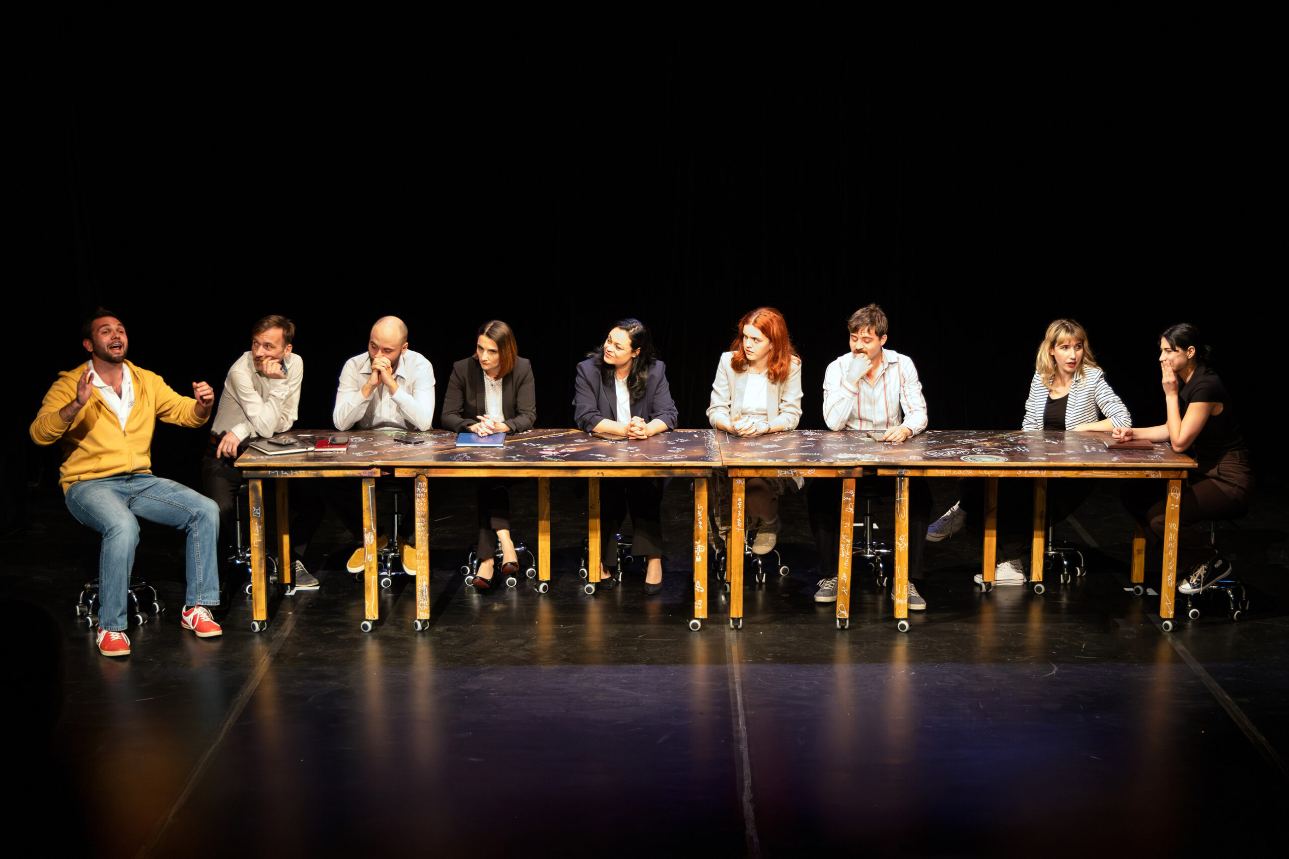 Panel of eight people seated at a long chalk-drawn table on a dark theater stage; the man in a yellow hoodie on the far left gestures while others listen.