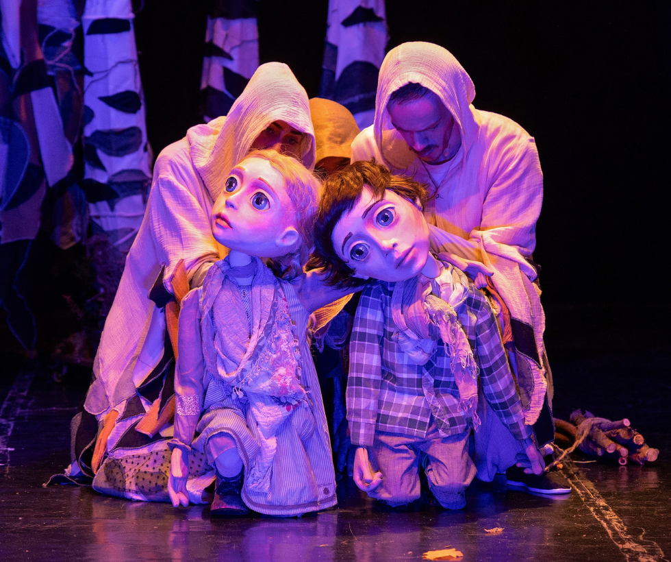 Puppet performance on stage: two oversized-eyed dolls kneel, surrounded by puppeteers in white robes under purple lighting.