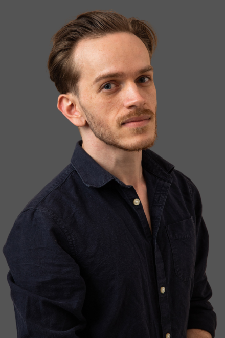 Portrait of a man wearing a dark blue button-down shirt, looking slightly to the right with a neutral expression against a gray backdrop.