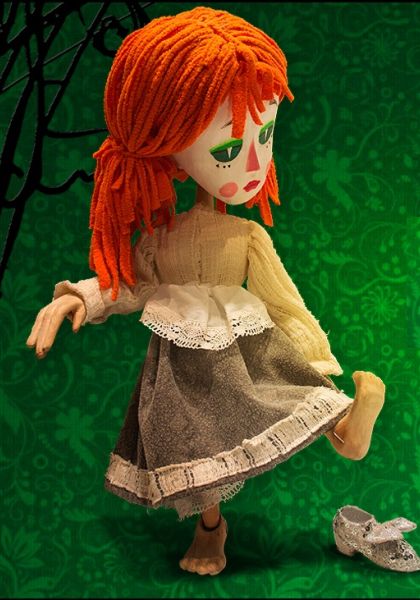 Soft fabric doll with bright orange yarn hair, beige sweater, gray lace-trimmed skirt, standing in a playful pose on a green patterned backdrop; a silver shoe lies to the side.