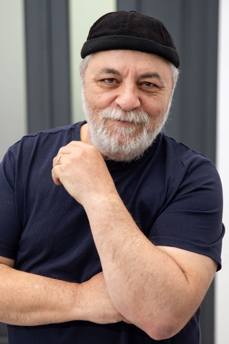 Older man with a gray beard wearing a black cap and navy t-shirt, arms crossed and a subtle smile.