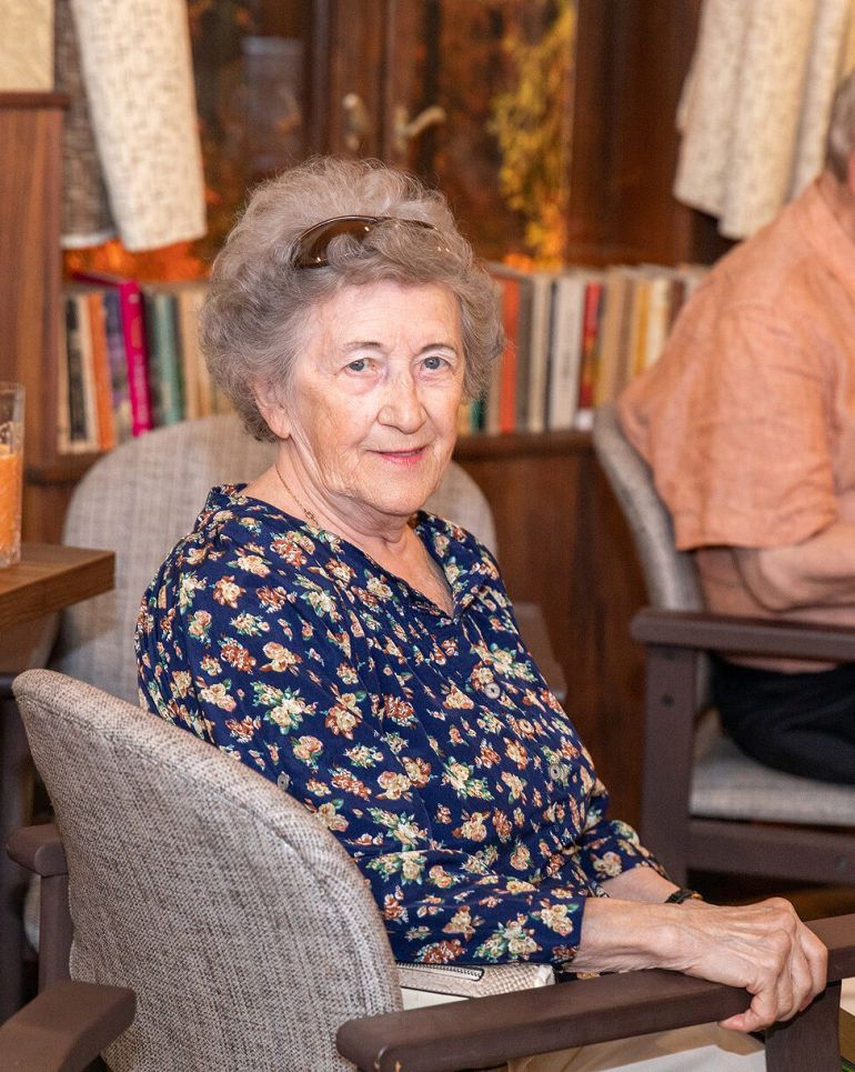 Smiling elderly woman in a navy floral blouse sits in a chair at a social gathering, with an orange juice drink on the table nearby.