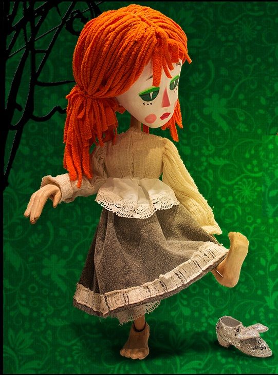 Soft fabric doll with bright orange yarn hair, beige sweater, gray lace-trimmed skirt, standing in a playful pose on a green patterned backdrop; a silver shoe lies to the side.