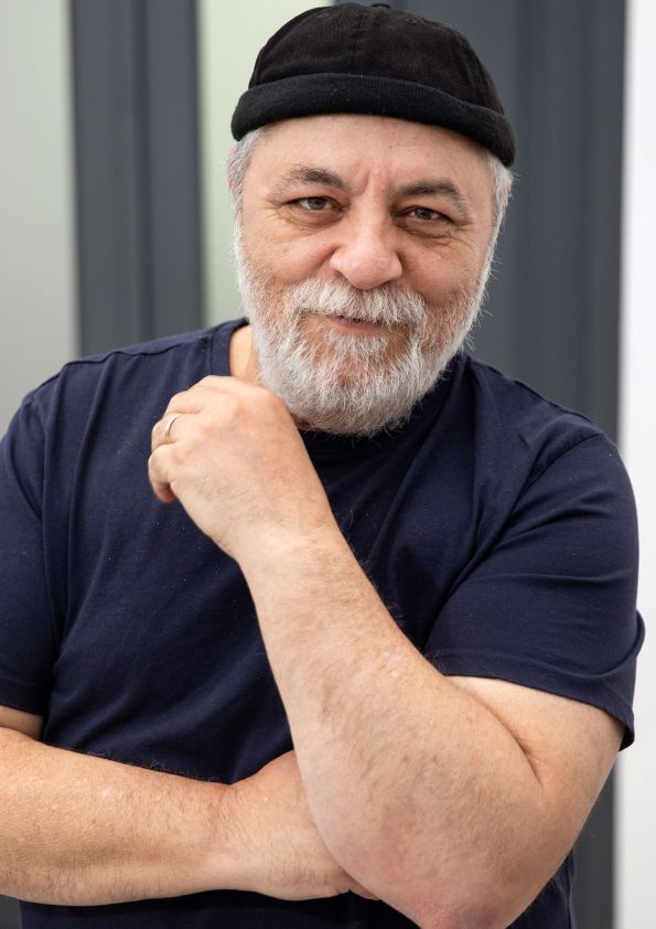 Older man with a gray beard wearing a black cap and navy t-shirt, arms crossed and a subtle smile.