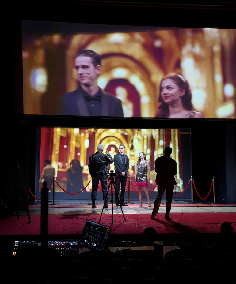Man in a tuxedo and a woman in a silver sequin dress stand on a red-carpeted stage with a gold, ornate backdrop; crew and equipment nearby.