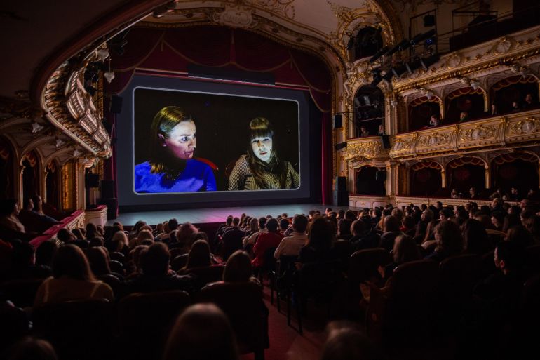Gilded theater interior with a large screen displaying two women in conversation, audience watching stage.