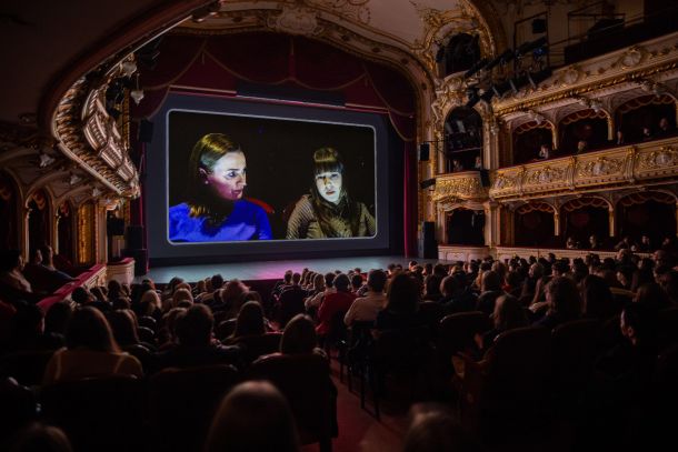 Gilded theater interior with a large screen displaying two women in conversation, audience watching stage.